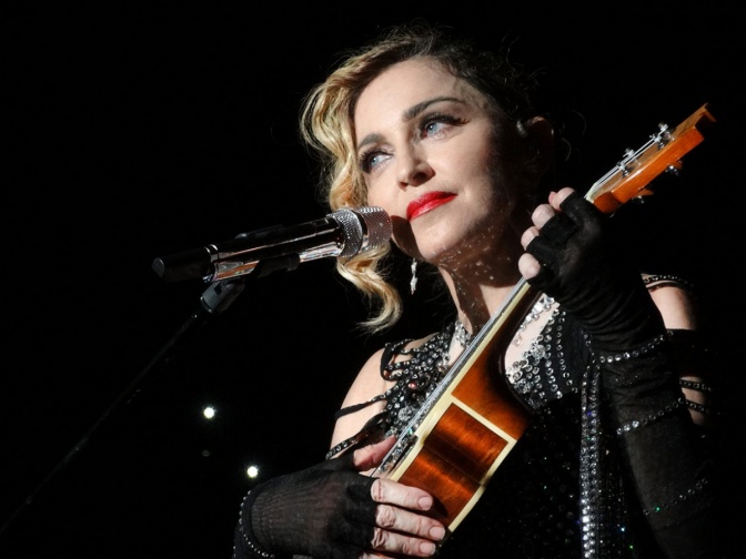 Madonna steht vor einem Mikrophon. Sie trägt schwarze Handschuhe ohne Finger und spielt auf einer Ukulele.