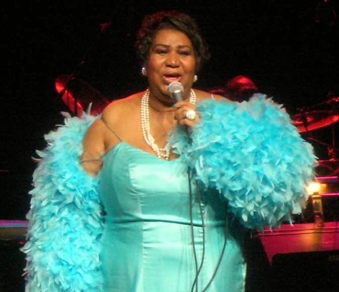 Aretha Franklin in einem Bühnenoutfit mit Federboa. Sie singt in ein Mikrophon. Ein Träger ihres Kleides ist über ihre Schulter gerutscht.