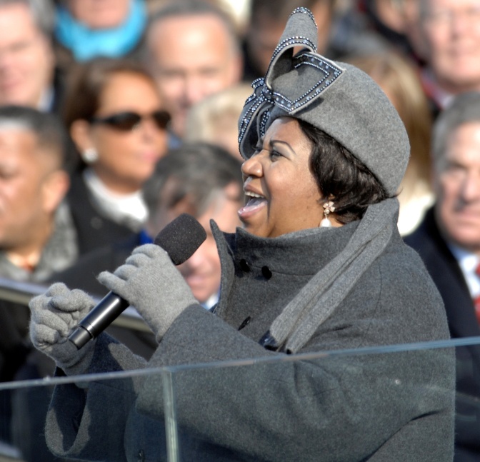 Aretha Franklin in einem grauen Mantel mit Mütze und Handschuhen. Sie singt in ein Mikroskop, hinter ihr sind verschiedene Menschen in einer Gruppe zu sehen, die ihr zuhören.