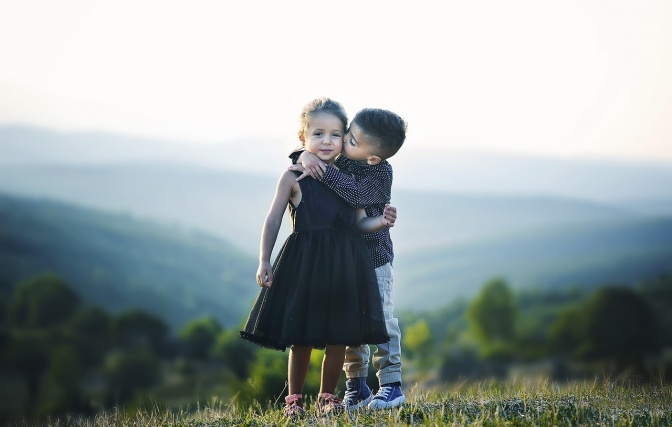 2 Kinder stehen vor einem Bergpanorama. Der Junge umarmt das Mädchen und küsst sie auf die Wange.