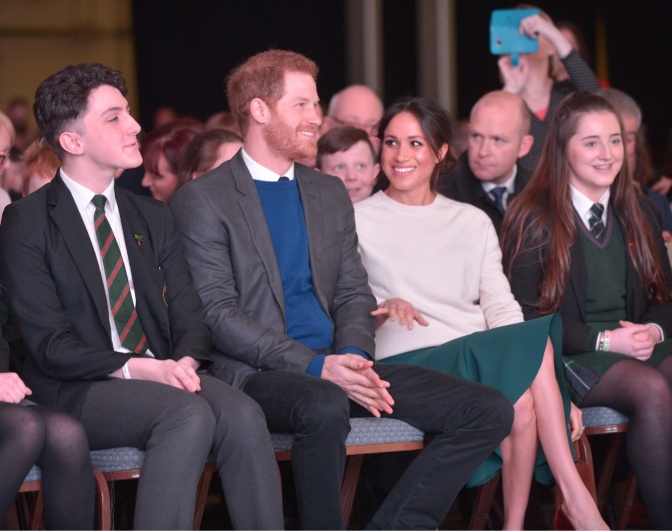 Prinz Harry und Herzogin Meghan sitzen auf zwei Stühlen in der ersten Reihe zwischen Schülern und Schülerinnen in Schuluniformen