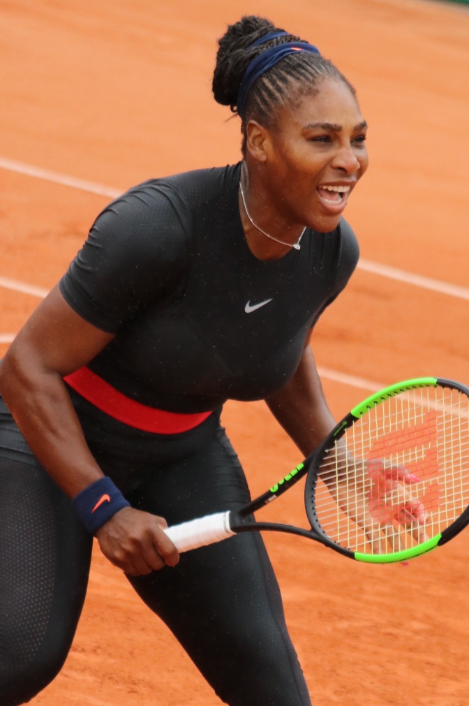 Serena Williams auf dem Tennisplatz. Sie hält einen Tennisschläger in der Hand. Die Haare hat sie zu kleinen Zöpfen geflochten und zurückgebunden. Sie lächelt triumphierend.