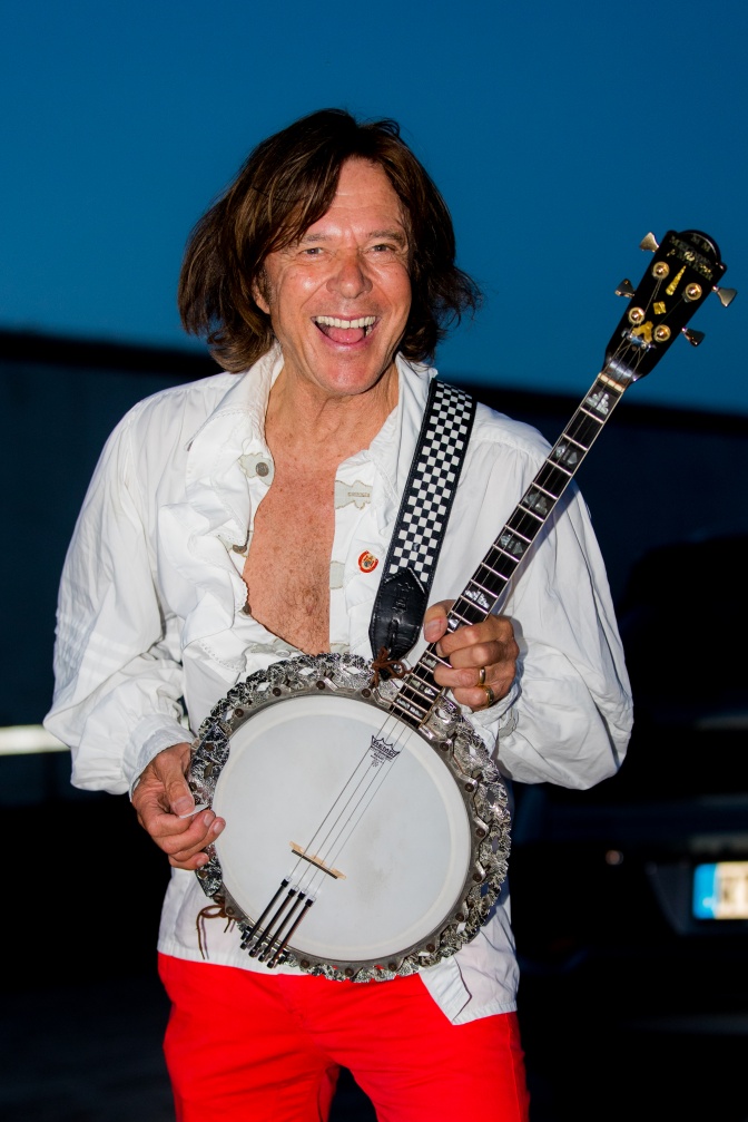 Jürgen Drews mit halblangen Haaren. Er trägt ein weißes, weit geöffnetes Hemd, durch das man seine nackte Brust sehen kann. Er hält ein Banjo in beiden Händen und lächelt ins Bild.