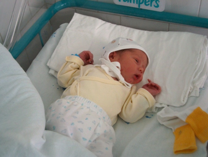 Ein neu-geborenes Baby in einem Babybett. Es trägt eine weiße Mütze und schläft.
