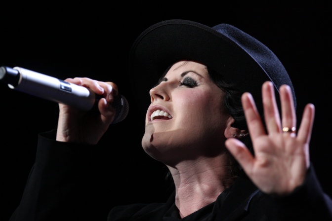 Dolores O'Riordan trägt einen schwarzen Hut. Sie singt mit geschlossenen Augen in ein Mikrophon.