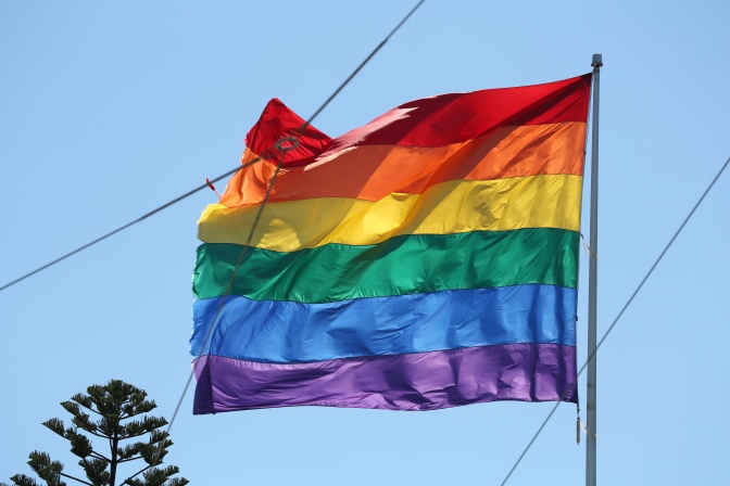 Eine Regebogenflagge weht vor blauem Himmel, im Hintergrund verläuft ein Stromkabel.