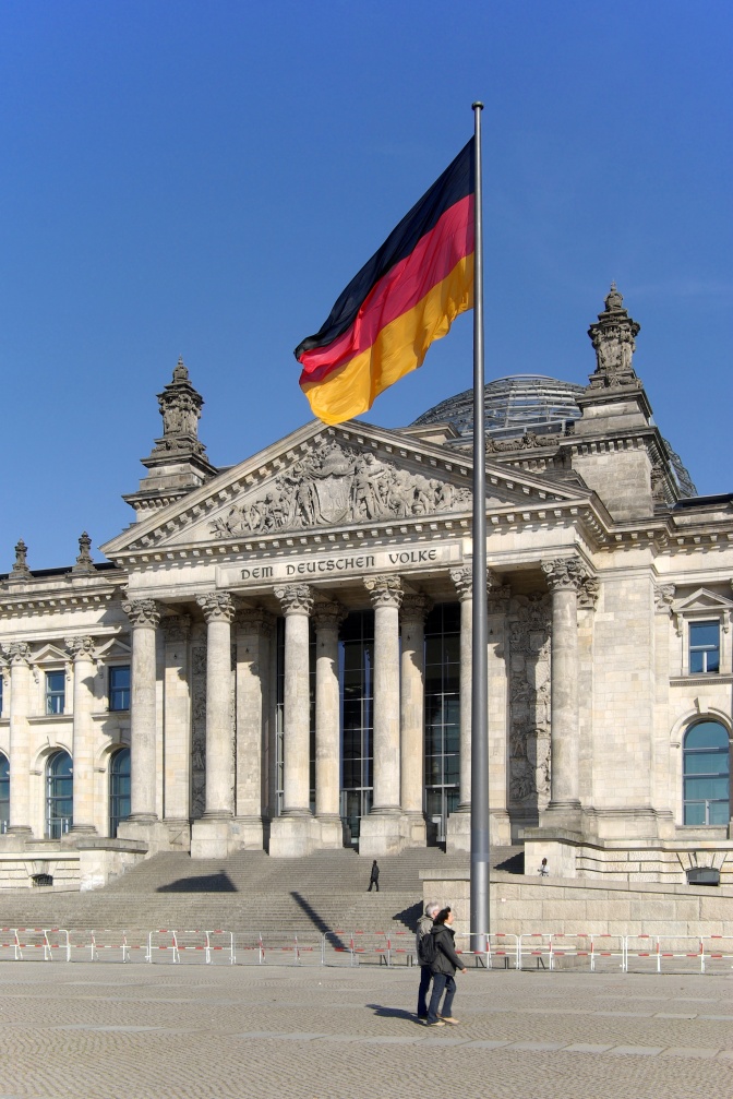 Das Reichstags-Gebäude in Berlin: Ein Bau mit mehreren Säulen. Davor weht die deutsche Flagge im Wind.