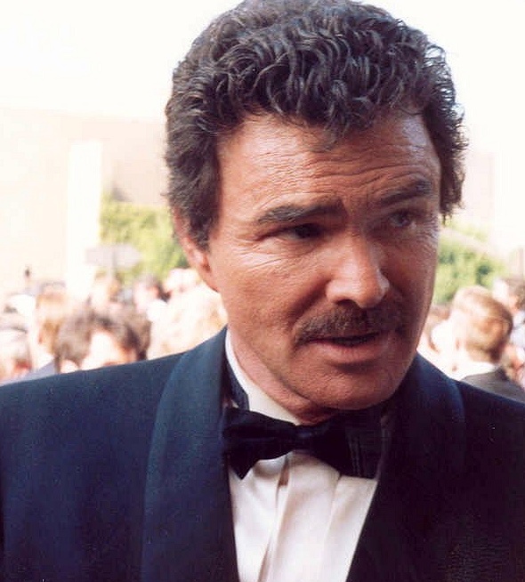 Burt Reynolds in Anzug und Fliege. Seine Haare sind kurz, braun und gelockt. Er trägt einen Schnäuzer.