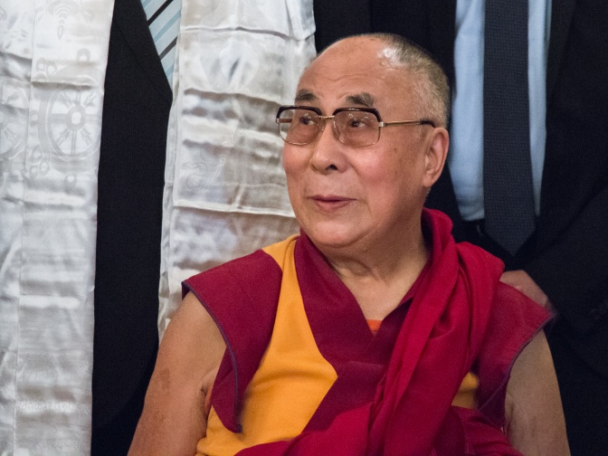 Der Dalai Lama trägt gewickelte Gewänder in dunkelrot und senfgelb. Er hat eine Glatze und trägt eine runde Brille.