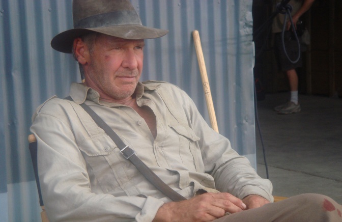 Harrison Ford trägt einen braunen Filzhut und ein helles Hemd. Er sitzt in einer Filmkulisse.