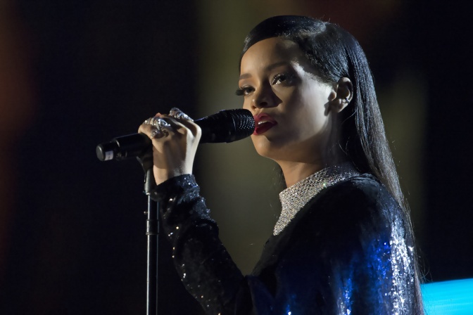 Rihanna singt auf der Bühne in ein Mikrophon. Si hat lange, geglättete schwarze Haare und trägt ein schwarzes Kleid.
