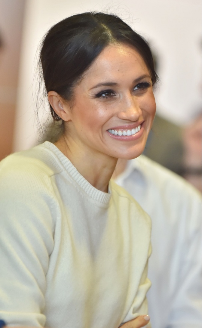 Herzogin Meghan von Sussex mit zurückgebundenen Haaren. Sie trägt einen hellen Pullover und lächelt.