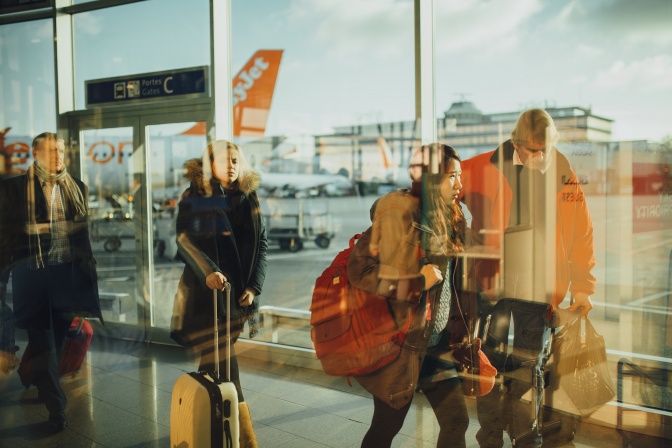 Mehrere Reisende betrachtet durch eine Glasscheibe. Sie tragen Gepäckstücke und laufen von rechts nach links.