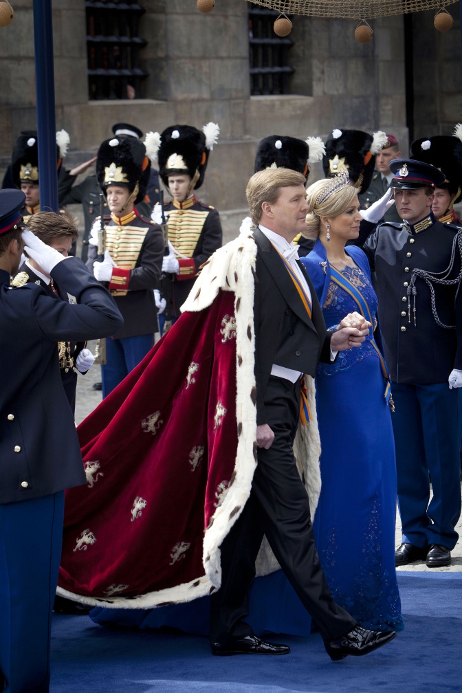 Màxima und Willem-Alexander der Niederlande schreiten nebeneinander. Er trägt einen roten Umhang mit Hermelin-Besatz, sie ein langes, königsblaues Kleid.