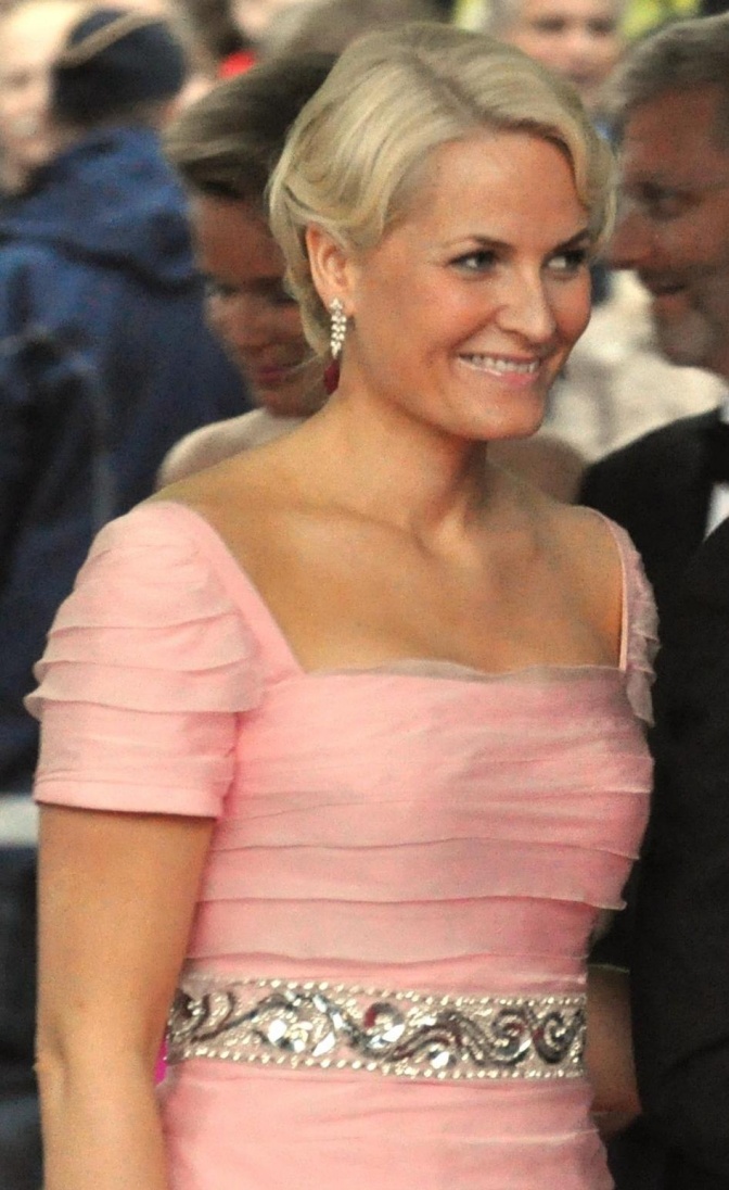 Mette-Marit von Norwegen in einem rosafarbenen Kleid mit kurzen Ärmeln und Glitzergürtel. Ihre blonden Haare hat sie hochgesteckt. Sie lächelt.