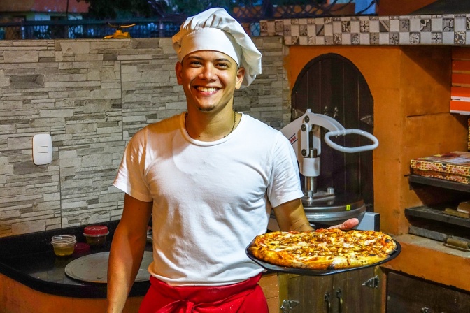 Ein Pizzabäcker trägt eine Kochmütze und ein weißes T-Shirt. Er hält eine fertig gebackene Pizza in der Hand und lächelt stolz. Im Hintergrund ist ein Steinofen zu sehen.