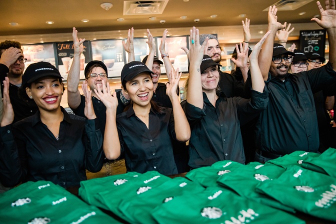 10 Personen tragen Starbucks Uniform, heben die Hände über den Kopf und bewegen die Hände mit gespreizten Fingern hin und her.