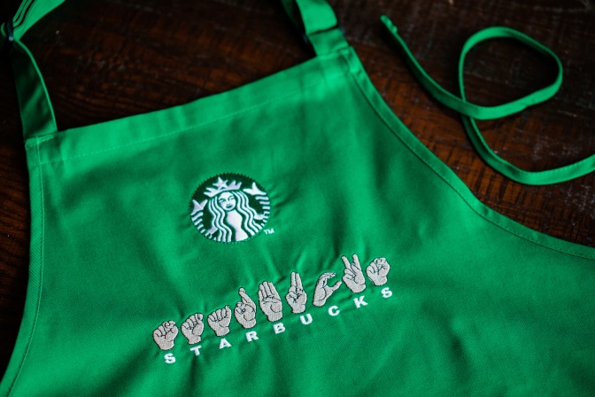Eine Schürze mit dem Starbucks-Schriftzug. Unter den Buchstaben des Schriftzugs STARBUCKS stehen die jeweiligen Gebärden für die einzelnen Buchstaben.