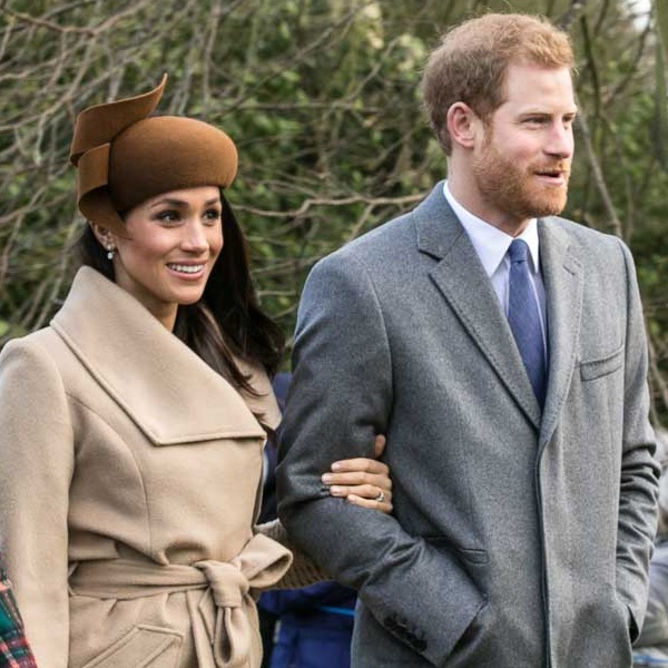 Prinz Harry und Herzogin Meghan in Winterkleidung. Sie hat sich bei ihm eingehakt. Beide lächeln.