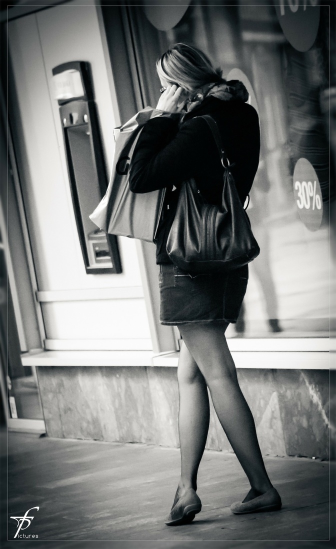 Ein schwarz-weiß-Foto einer Frau, die im Gehen telefoniert. Sie trägt einen Minirock und eine große Handtasche.