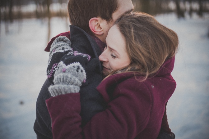 2 Menschen umarmen sich, ein Mann und eine Frau. Beide tragen dicke Winterkleidung. Im Hintergrund sieht man eine Winterlandschaft.