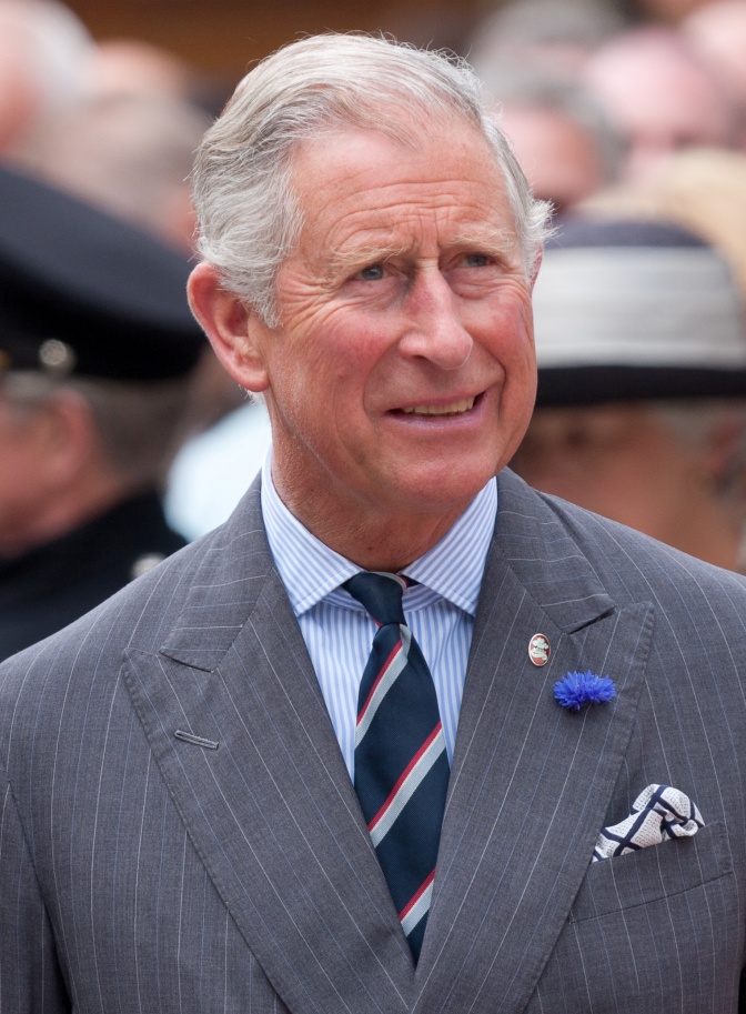 Prinz Charles mit nach hinten frisierten grauen Haaren. Er trägt Anzug und Krawatte. Am Revers hat er eine blaue Nelke angesteckt.