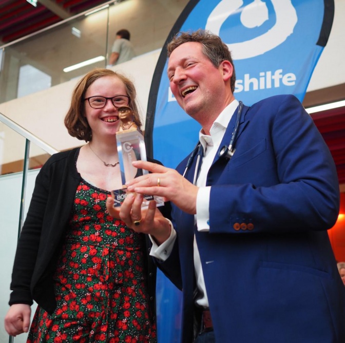 Natalie Dedreux und Eckart von Hirschhausen stehen nebeneinander. Er hält den dem Preis BOBBY in der Hand. Im Hintergrund sieht man das Logo der Lebenshilfe.