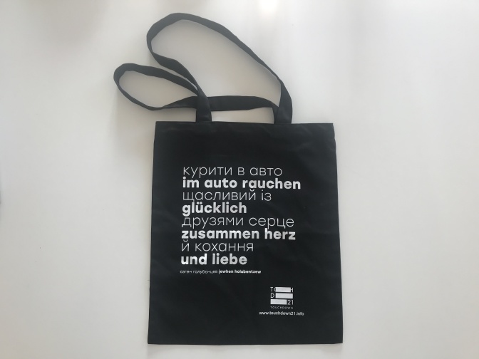 Eine schwarze Stofftasche mit weißer Schrift. Aufgedruckt ist ein zweisprachiges Zitat in deutsch und ukrainisch.