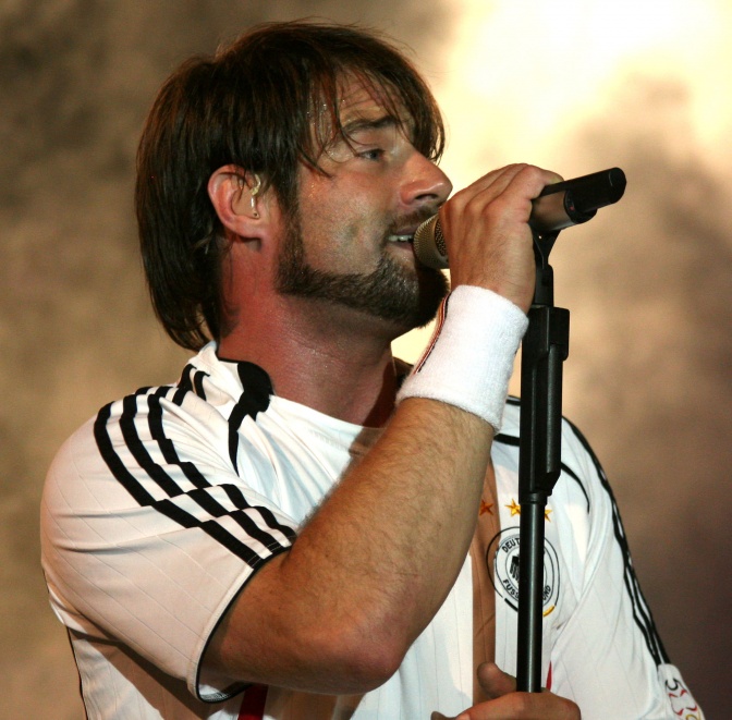 Sasha in einem Trikot von Adidas mit 3 Streifen auf der Schulter. Er hat längere Haare und einen Bart. Er singt in ein Mikrophon.