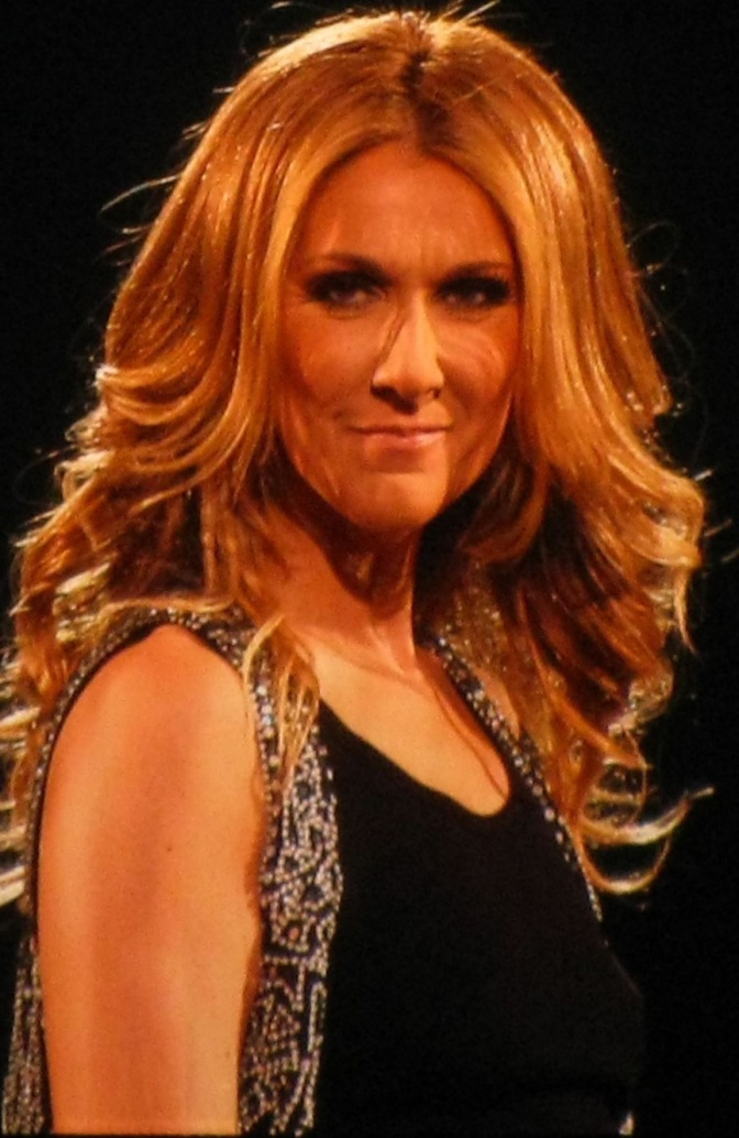 Céline Dion in einem schwarzen Kleid mit Trägern und mit gewellten, blonden Haaren. Sie dreht sich seitlich der Kamera zu.