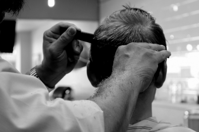 Ein Friseur schneidet einem Mann einen Kurzhaarschnitt. Er fährt mit einem Kamm durch die Haare und schneidet an der Schere entlang.