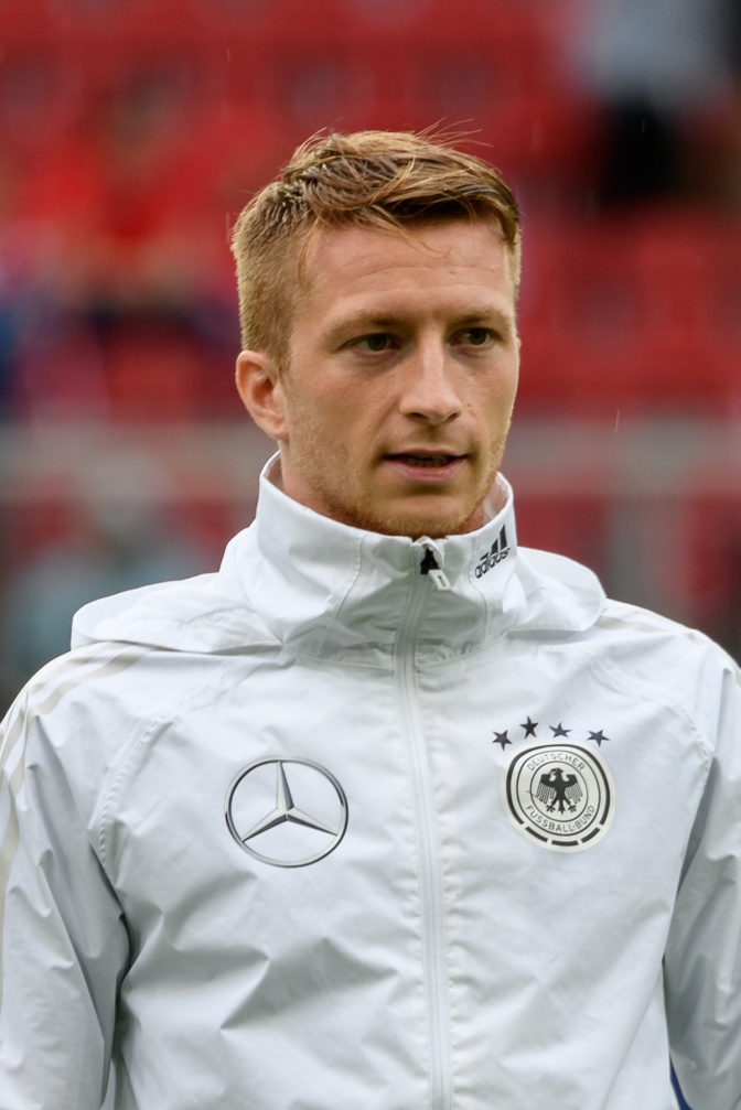 Marco Reus in einer weißen Trainingsjacke der deutschen Nationalmannschaft. Im Hintergrund sieht man die Bestuhlung des Stadions.