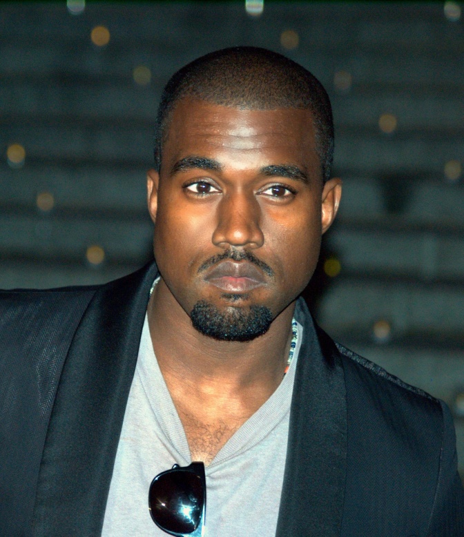 Kanye West in einem grauen Shirt und mit Sakko. Er hat eine Sonnenbrille in den Bund des T-Shirts geklemmt. Er trägt einen kurzrasierten schwarzen Bart und auch seine Haare sind sehr kurz.