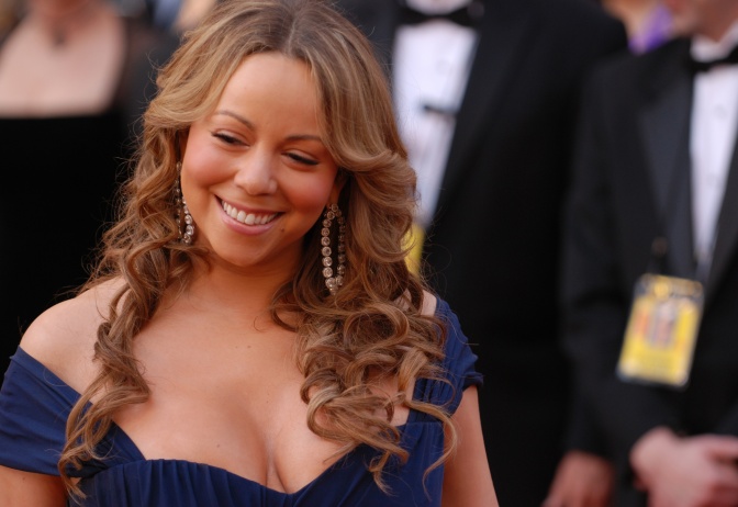 Mariah Carey in einem tief ausgeschnittenen dunkelblaues Kleid, mit eingedrehten Haaren und großen Ohrringen. Sie lächelt. Im Hintergrund sieht man Männer in Anzügen.