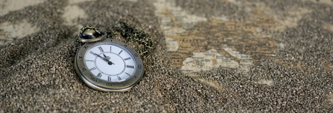 Eine Taschenuhr liegt im Sand. Unter dem Sand erkennt man Teile einer Weltkarte.