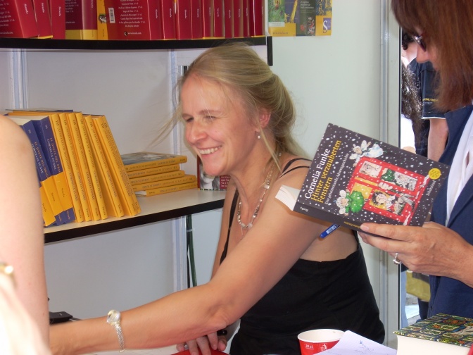 Cornelia Funke sitzt in einer Bücherei und signiert Bücher. Sie lächelt. Neben ihr steht ein Kind mit einem aufgeschlagenen Buch in der Hand.