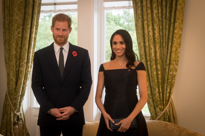Prinz Harry und Herzogin Meghan stehen in eleganter Kleidung vor einem Sprossenfenster mit schweren Gardinen.