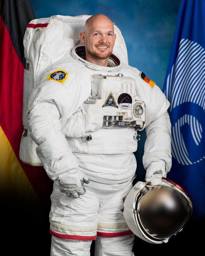 Alexander Gerst in einem Weltraumanzug. Den runden, durchsichtigen Helm hält er in der Hand. Er hat eine Glatze und lächelt.