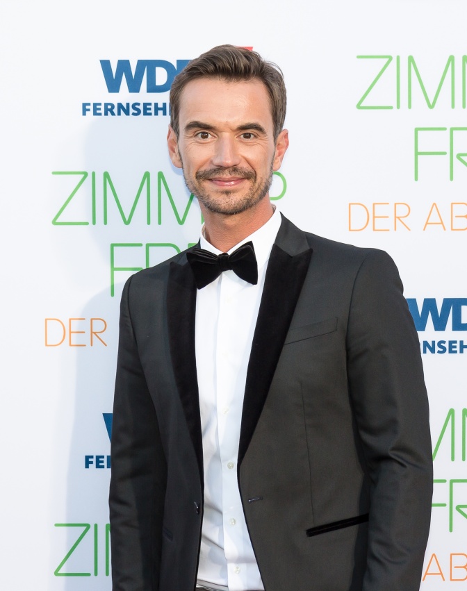 Florian Silbereisen in Anzug, Hemd und Fliege vor einer Logowand, auf der Zimmer frei steht. Er lächelt.