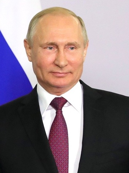 Wladimir Putin in Anzug und Krawatte. Er steht vor einer russischen Flagge.