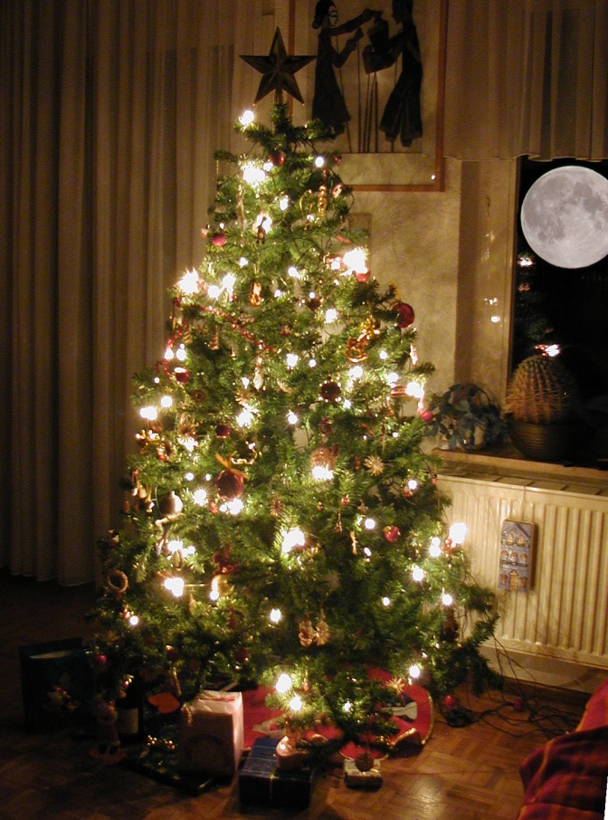 Ein geschmückter Weihnachtsbaum steht auf Parkettboden. An der Wand dahinter hängt ein Kunstdruck. Vor dem Fenster sieht man den Vollmond.