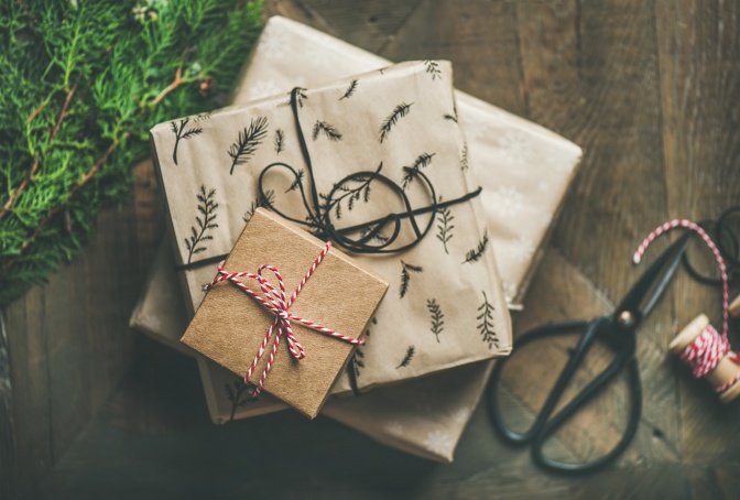 Mehrere Geschenkpäckchen mit Schleifen in verschiedenen Farben liegen in einem Stapel übereinander. Daneben liegen ein Tannenzweig, Geschenkband und eine Schere.