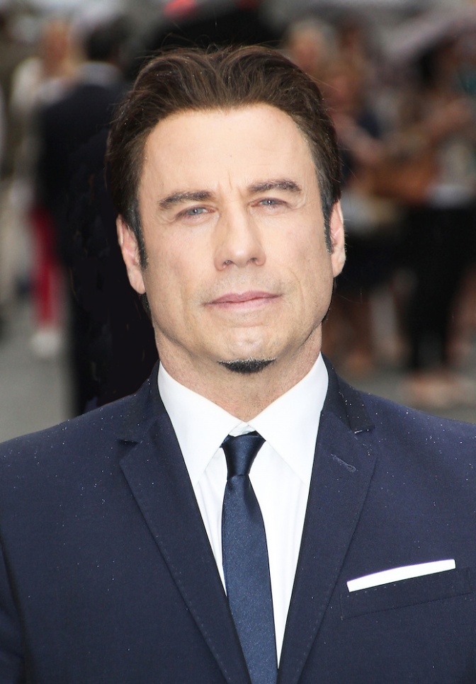John Travolta in Anzug und Krawatte mit vollem, braunem, nach hinten frisiertem Haar und einem kleinen Kinnbart.