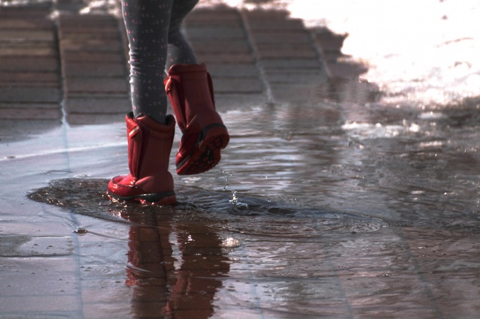 Ein Paar rote Stiefel springen in eine Pfütze auf Pflastersteinen. Das Wasser spritzt nach oben.