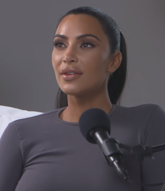 Kim Kardashian sitzt vor einem Mikrophon und spricht hinein. Sie hat einen streng nach hinten frisierten Pferdeschwanz und trägt ein schlichtes Langarm-Shirt.