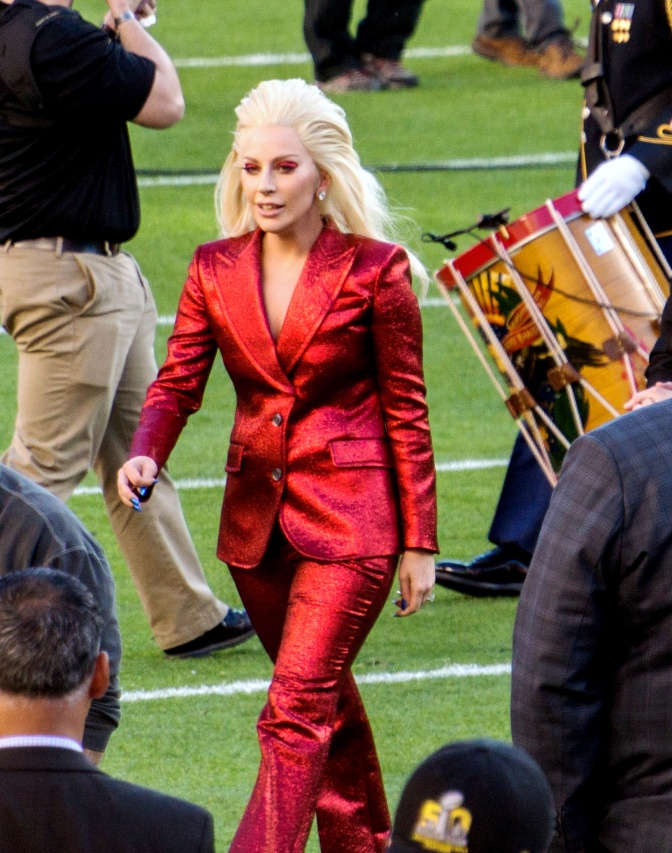 Lady Gaga läuft über einen Rasen. Sie hat lange weißblonde Haare und trägt einen Anzug. Um sie herum laufen andere Menschen, einer davon hat eine Trommel umhängen.