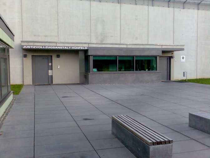 Ein Betonbau mit einer schweren Metalltür. Über den Eingang steht in Großbuchstaben Justizvollzugsanstalt Hünfeld. Im Vordergrund des Bildes ist eine Bank ohne Lehne zu sehen.