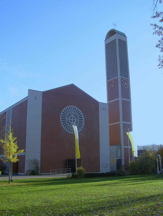 Eine Kirche in Backsteinoptik mit einem Glockenturm. Sie steht auf einer grünen Wiese vor blauem Himmel. Sie hat ein rundes Fenster, in dessen Mitte ein Kreuz zu sehen ist.