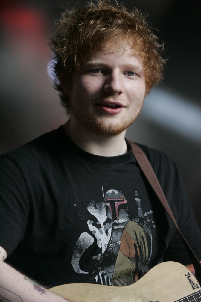 Ed Sheeran in einem schwarzen T-Shirt mit Print. Er hat eine akustische Gitarre an einem Lederband umhängen.
