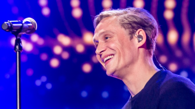 Matthias Schweighöfer hat einen Kopfhörer im Ohr und sitzt lächelnd vor einem Mikrophon. Im Hintergrund sind verschiedenfarbige Scheinwerfer zu sehen.